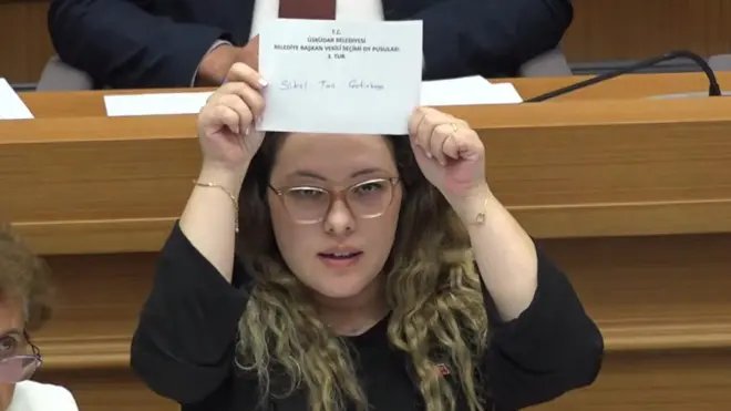 Üzerinde siyah kıyafet olan, gözlüklü ve saçlarının ucu sarı bir belediye çalışanı oy pusulasını meclise gösteriyor. Pusulanın üzerinde CHP'nin adayı Sibel Can Çetinkaya'nın adı yazıyor.