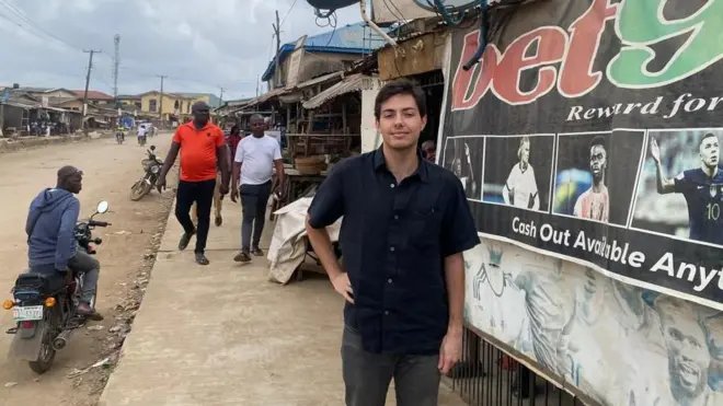 Koyu saçlı genç bir İspanyol olan Carlos, yol kenarındaki bir binanın yanında, üzerinde büyük bir spor bahis reklamı bulunan bir tabelanın yanında duruyor. Tabelada dikkat çekici bir şekilde "bet9" ve "İstediğiniz Zaman Para Çekme İmkanı" yazıları ile futbolcuların resimleri yer alıyor. Koyu renk kısa kollu bir gömlek ve koyu renk pantolon giymiş. Nijerya'da, asfaltlanmamış bir yolun yanında, arkasında yayalar ve bir motosikletli ile tabelanın üzerinde iki büyük uydu anteni bulunan bir kentsel ortamda bulunuyor.