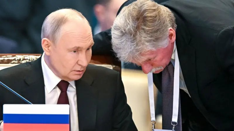 Putin Rus bayrağı görünen bir massada oturuyor, Peskov yanına eğilmiş, boynunda konferans kimliği sarkıyor, Putin'in sözlerini dinliyor gibi görünüyor.