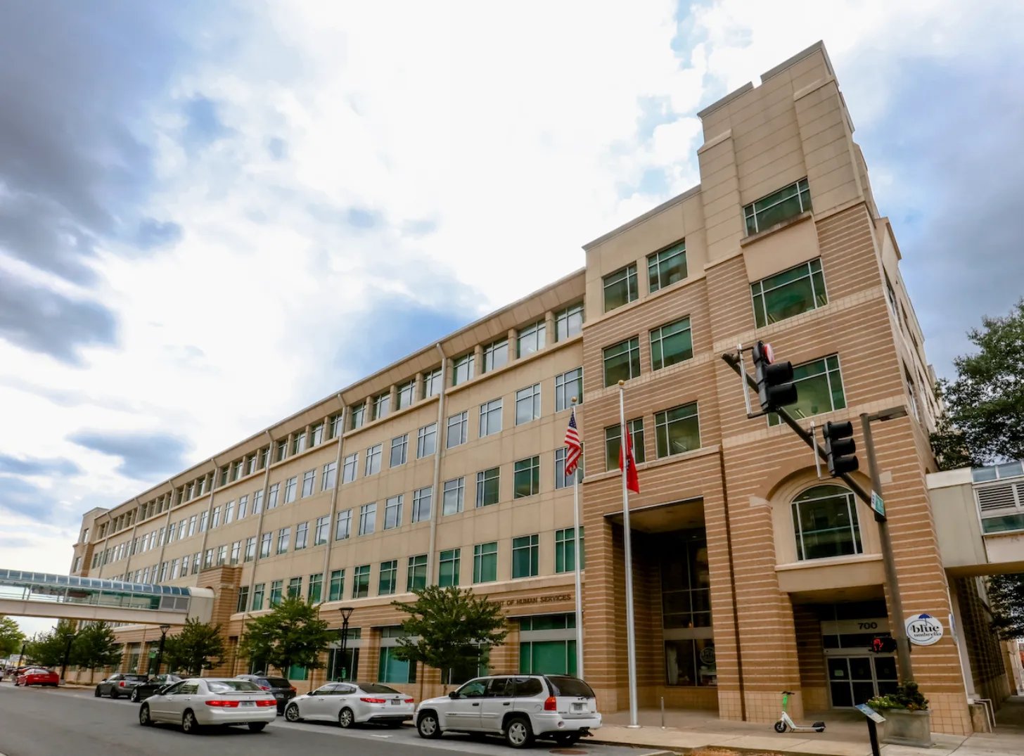 Brick and glass mid-rise building with multiple floors and windows, American flag and red flag hanging near entrance, parked cars in front, traffic light visible, trees and cloudy sky.