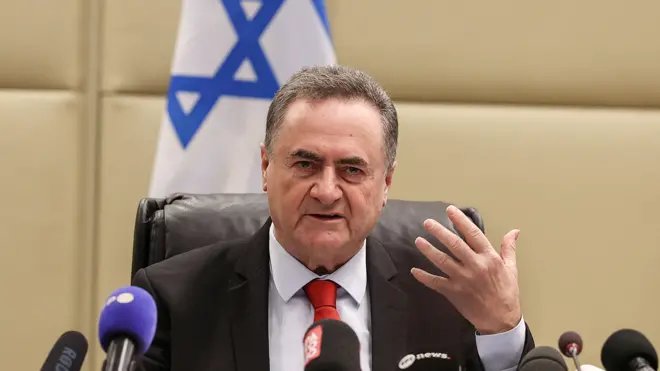 Israel Katz üzerinde bir takım elbiseyle bir masada oturmuş, yüzünde gergin bir ifadeyle konuşurken elini kaldırıyor. Önünde mikrofonlar, arkasında ise bir İsrail bayrağı var