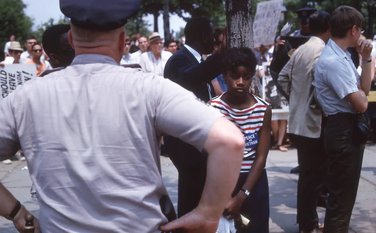View from behind of a police officer with arms on hips looking out at crowd of demonstrators