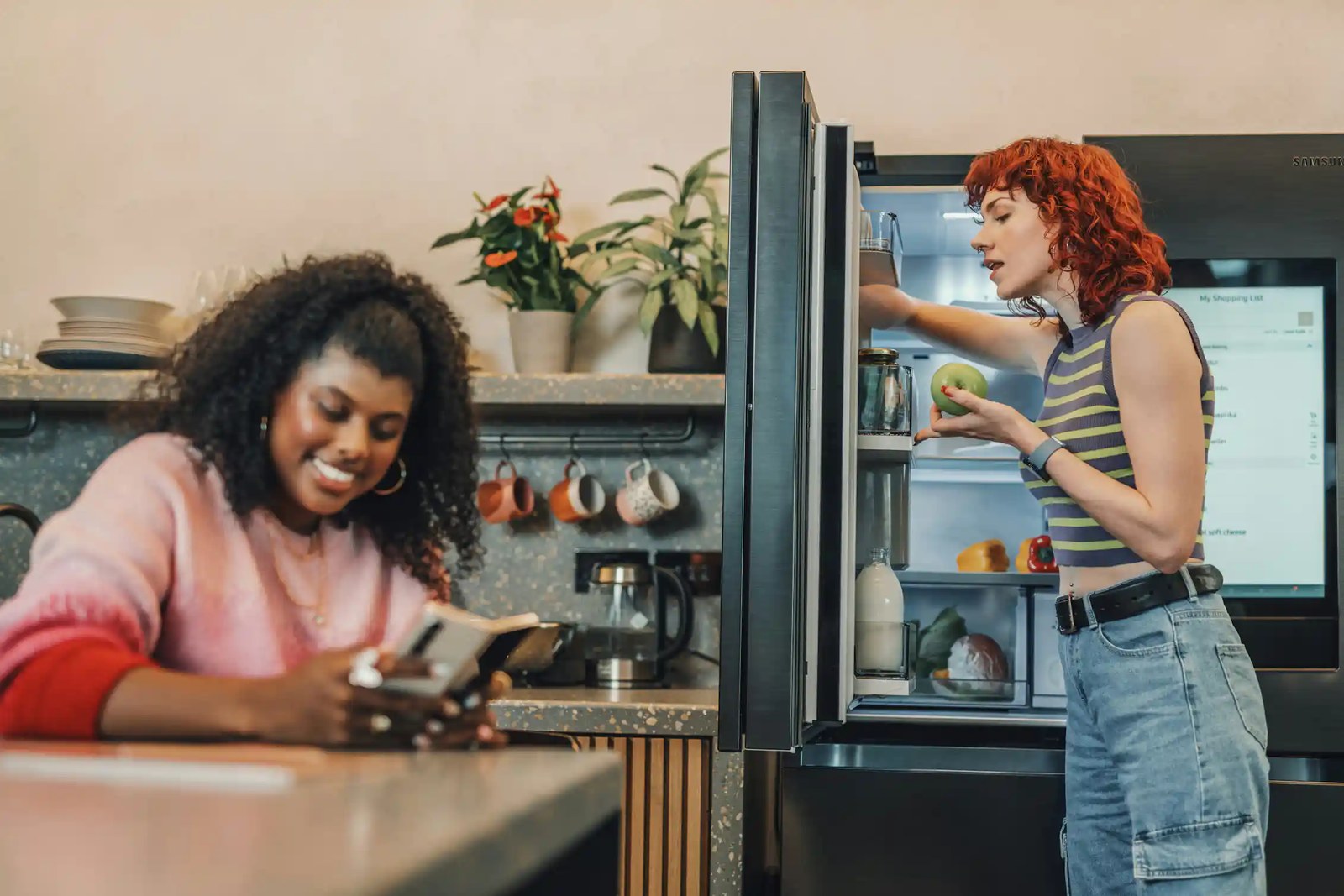 A woman sits at a table, scrolling on her phone and smiling, while another woman looks through a fridge while speaking to her