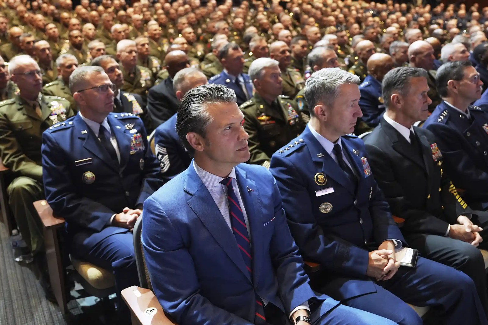 A man in a blue suit sits in an auditorium, with uniformed officers filling the seats beside and behind him.