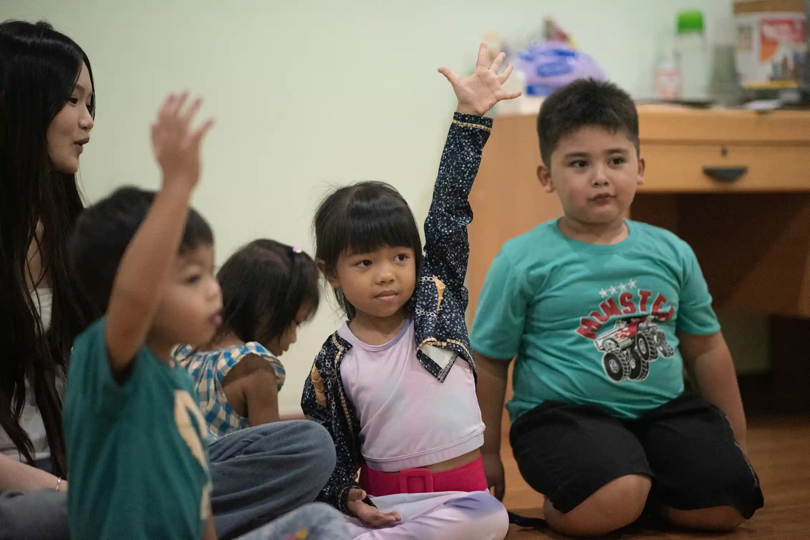 Children sitting in a circle, many looking attentive with their hands up.