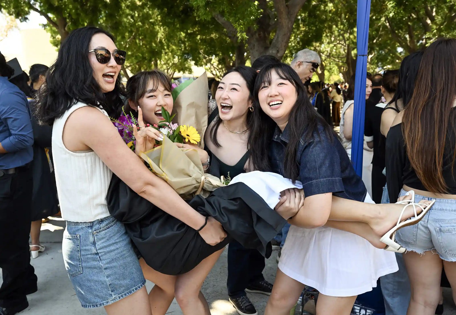 Four young women smile and pose as they lift one of the women, wearing a black graduation gown and carrying flowers, in the air.