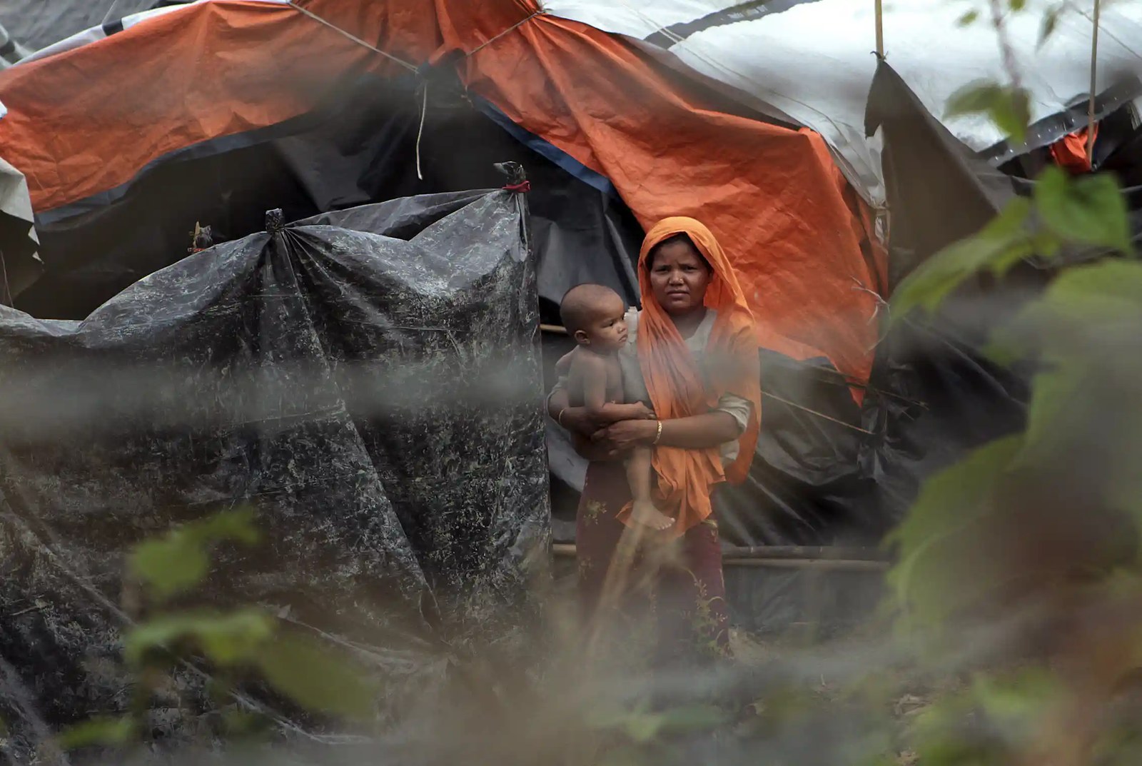 A woman holds a baby and looks through a chain-link fence.