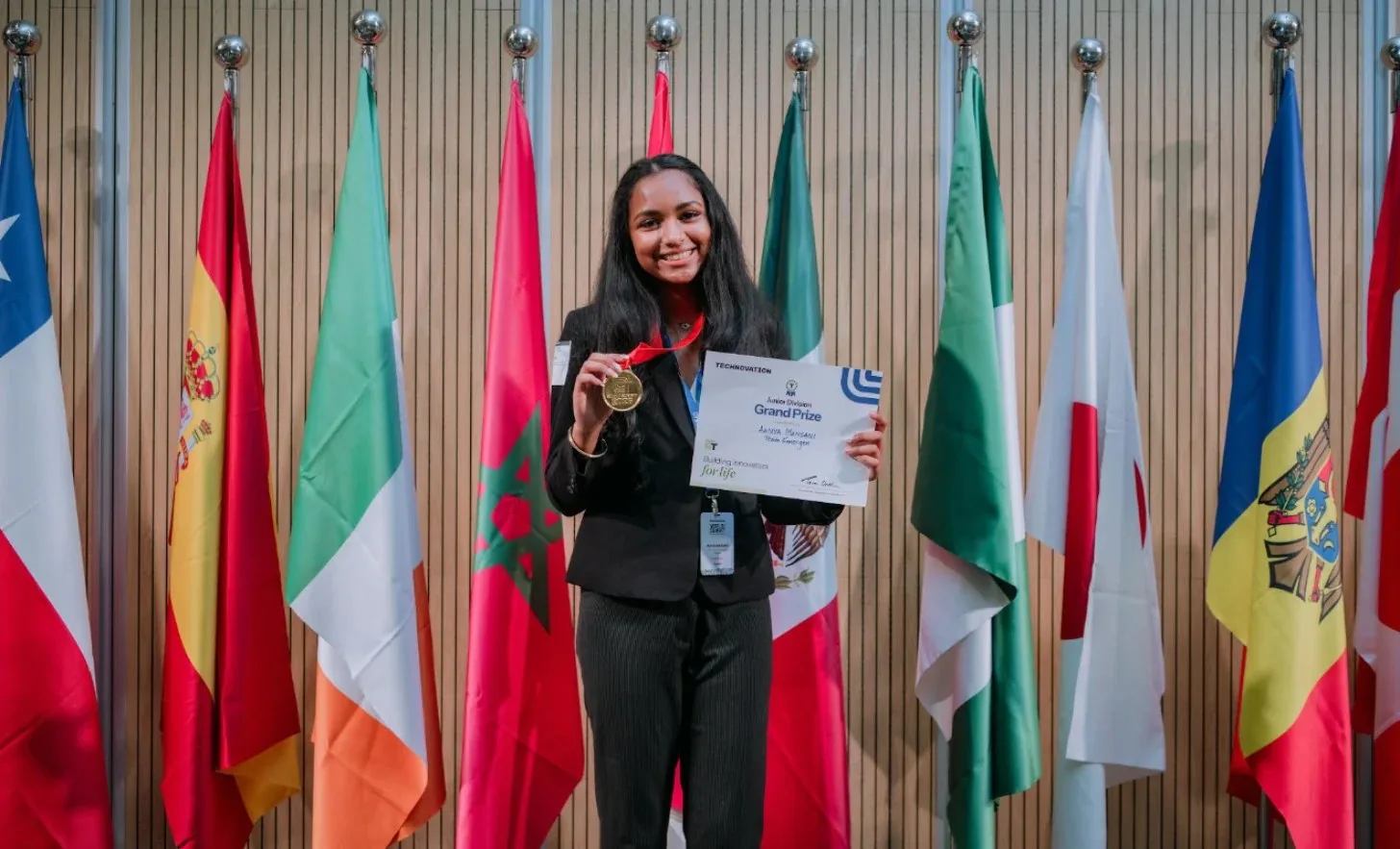 Young teen girl in black blazer holding a gold medal and certificate labeled "Grand Prix" stands in front of a backdrop of international flags.