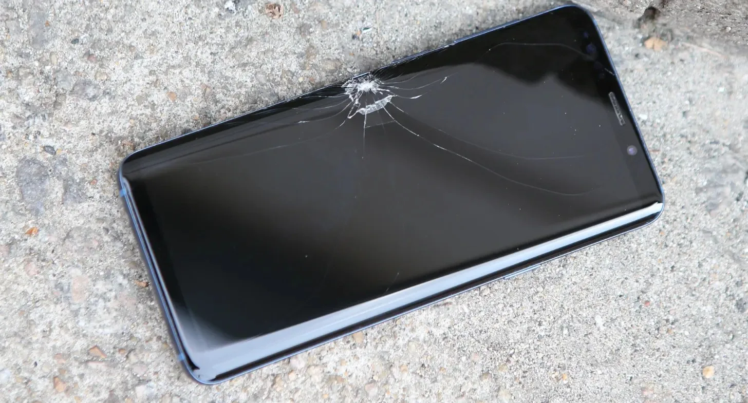 A smartphone with a cracked screen lies on a concrete surface. The display is dark, with visible damage radiating from the upper left corner.