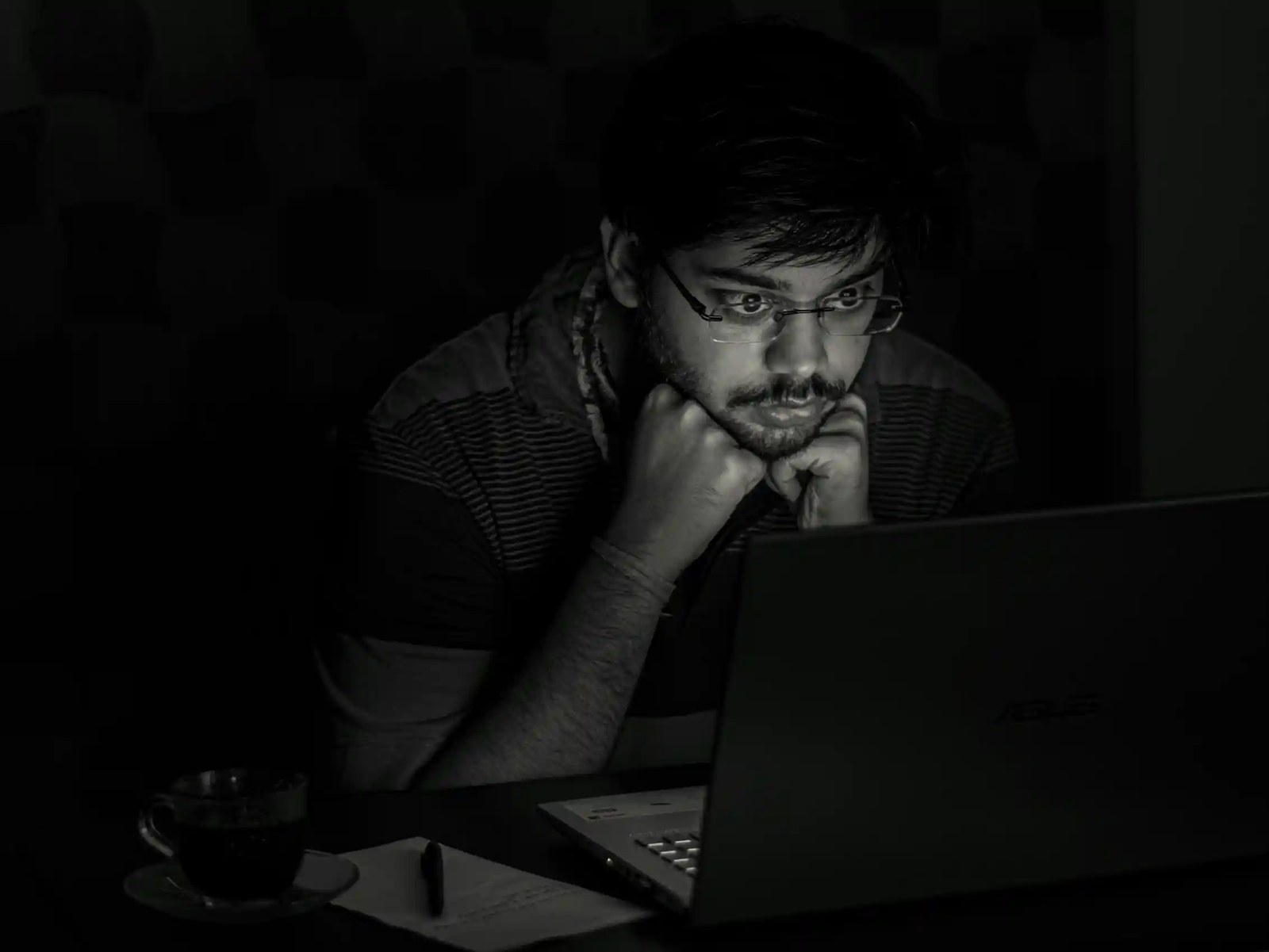 Black and white photo of a man in dark room, his face illuminated by the glow of a computer screen