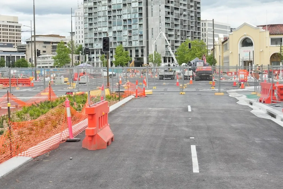 Road blocked off by fences and orange road blocks. A crane in the distance and buildings in the distance.
