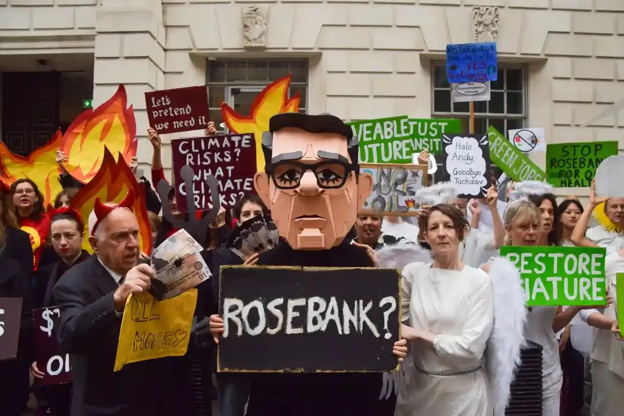 A protester in a PM mask holds a ‘Rosebank?’ sign amid activists dressed as devils and angels