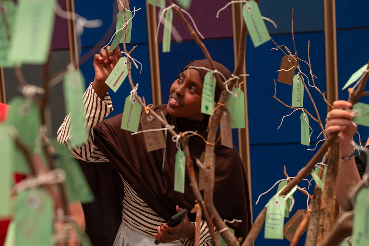 A teenage girl in a hijab reads tags tied to tree branches.