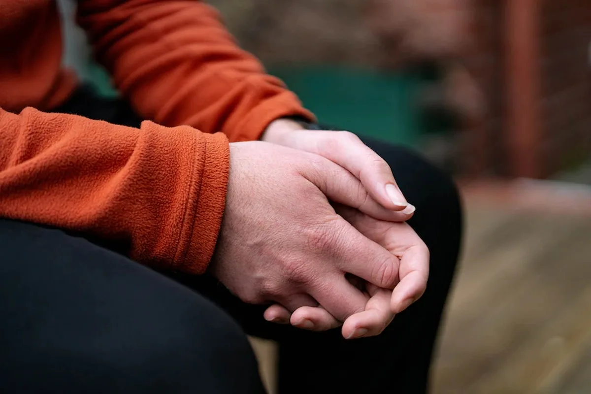 Close up of the hands of a man, wearing an orange jacket, clasped together as he sits in a backyard on a wooden deck.