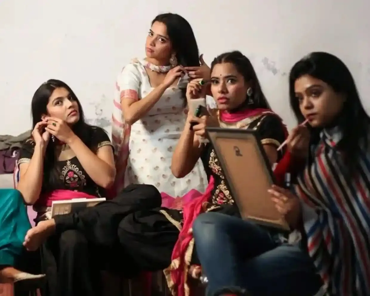 Four young women sitting side by side, applying makeup and doing their hair. 