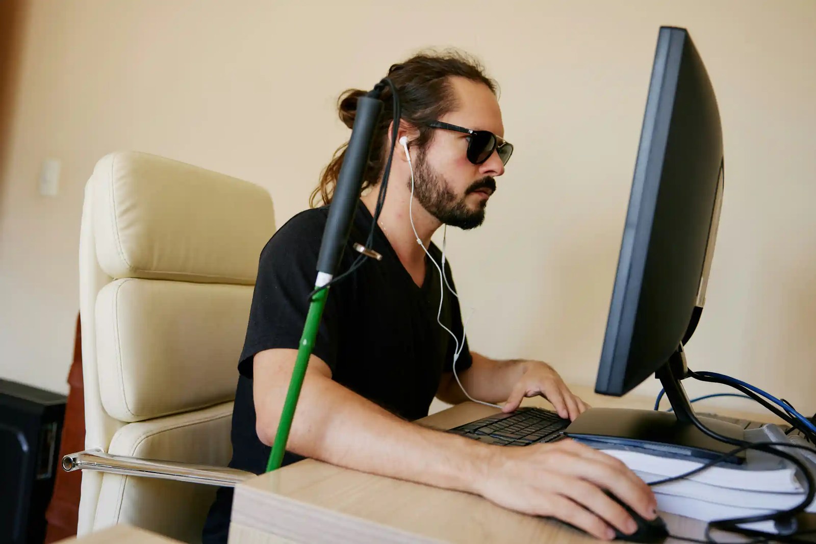 A person wearing sunglasses and wireless headphones works on a computer. A white cane rests beside him