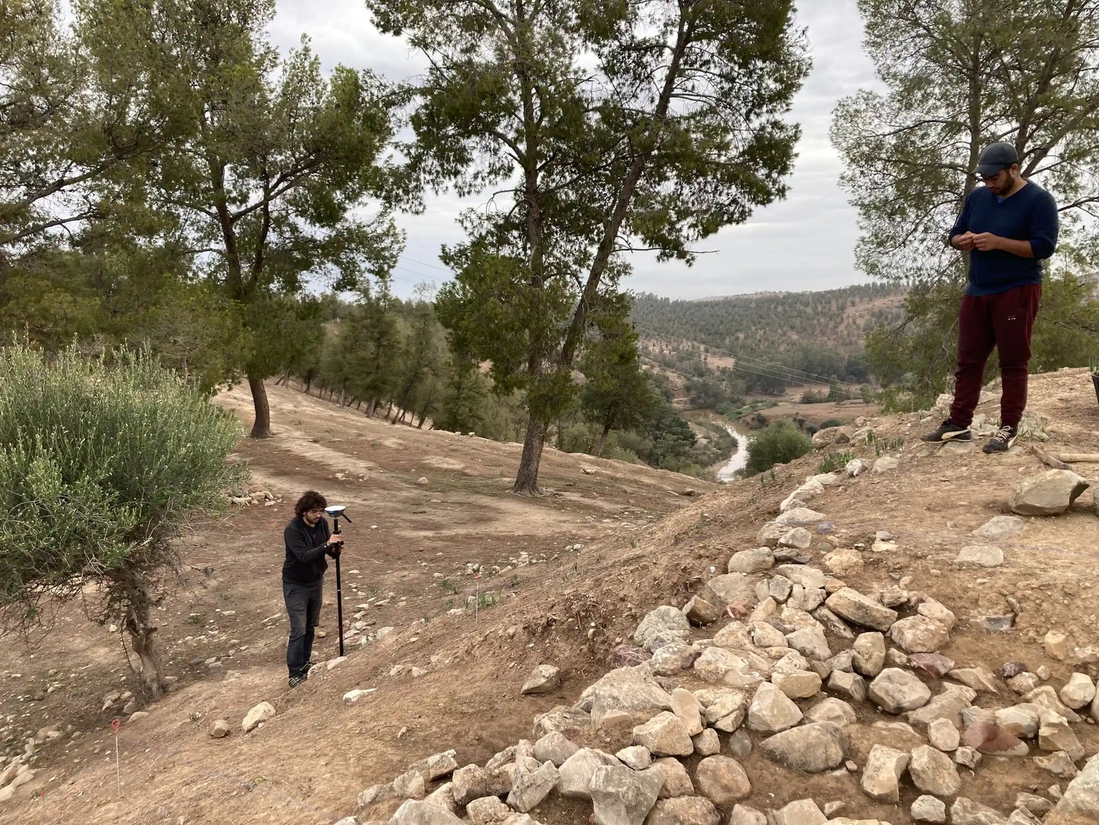 A man stands with a device on a tripod pointed at stacks of stones forming a ridge in the ground, another man standing above it, both dressed in black.