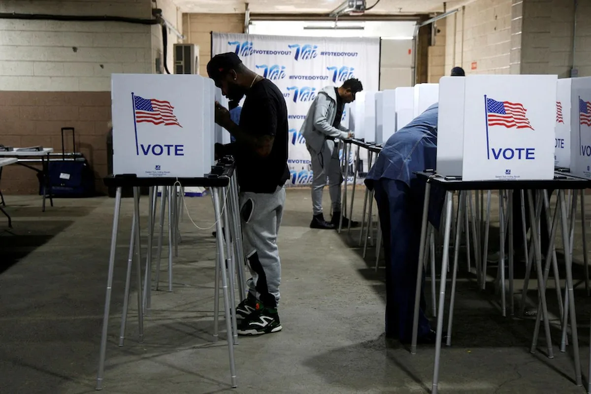 A room set up as a voting booth, people standing in sections separated by cardboard walls that say VOTE with the US flag