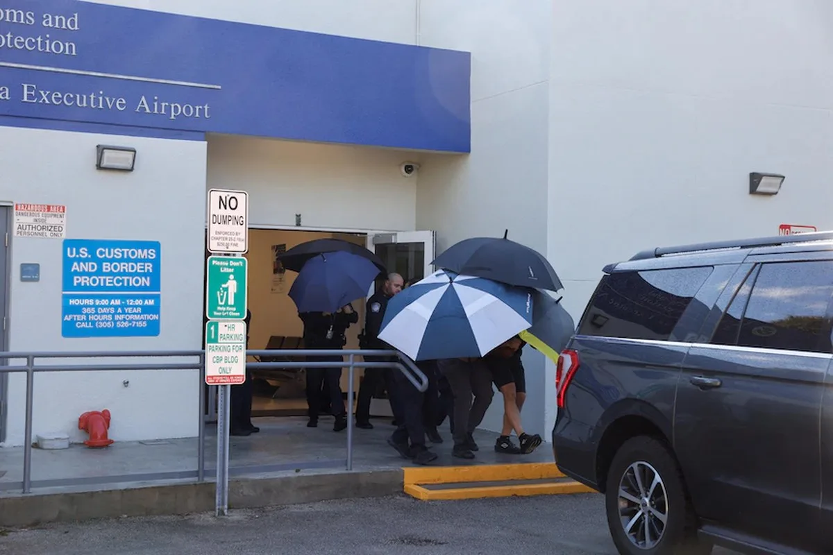 A man is shielded from view by multiple people holding umbrellas as he walks between an airport exit door and a car.
