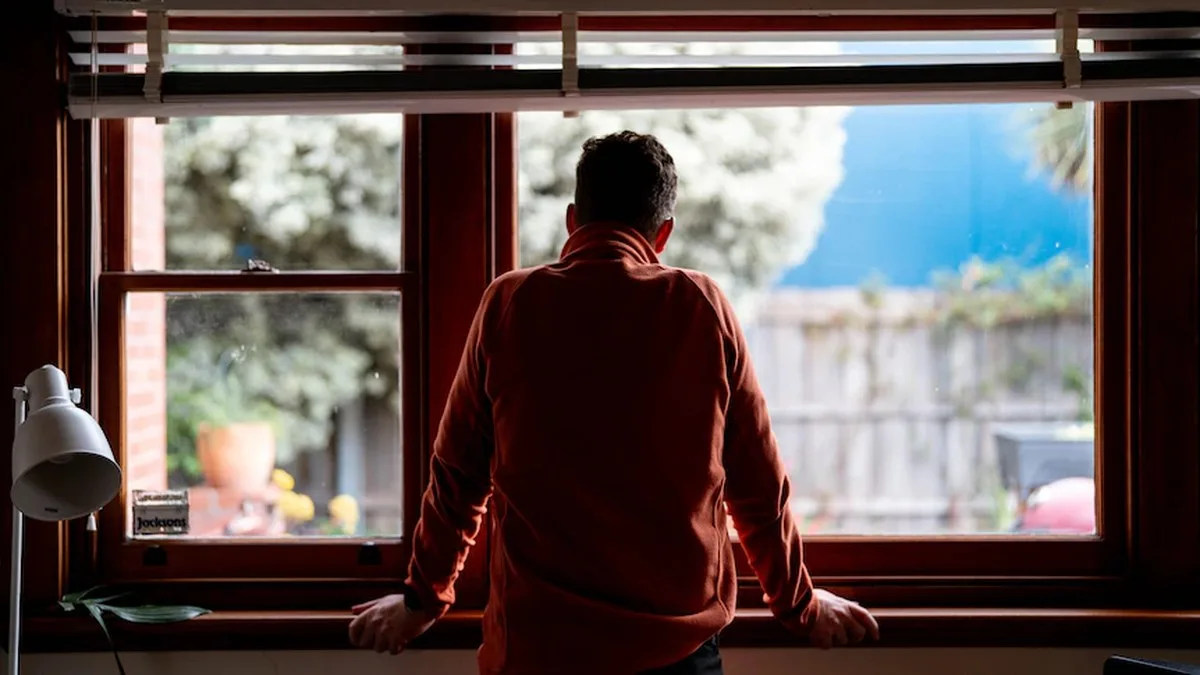A man, wearing an orange jacket, looking out the window in his dining room.