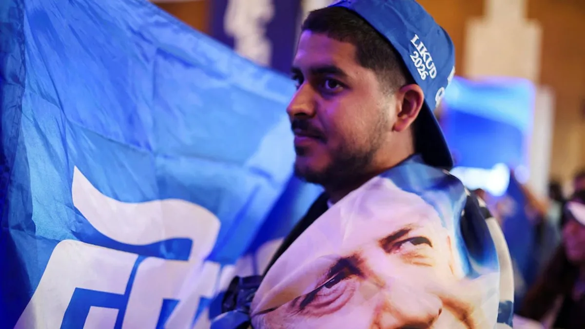 A man wraps a flag with Benjamin Netanyahu's face on it around his body and wears a blue hat that says Likud 2026.