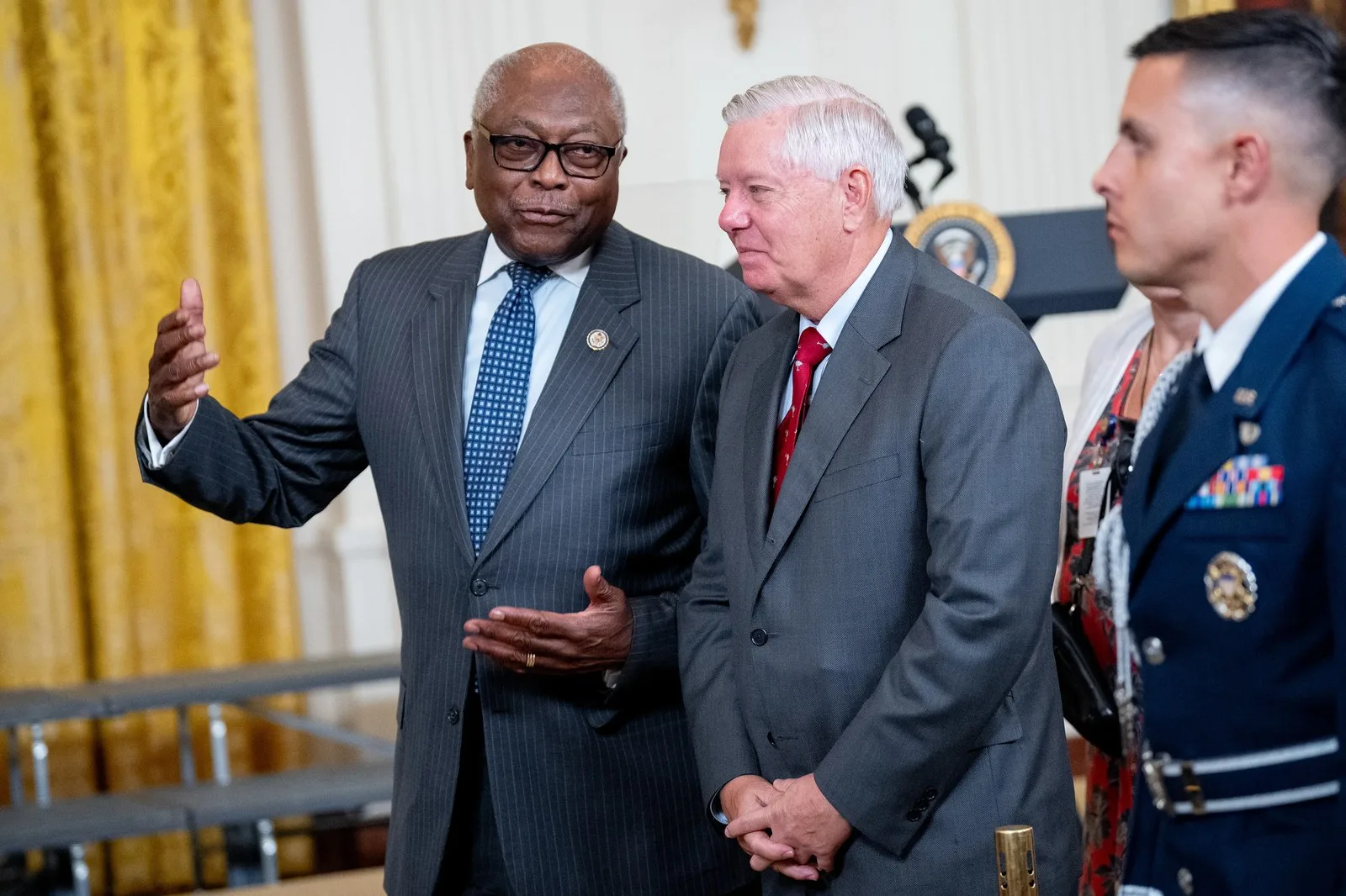 Two political figures visit during an event at the White House. The individual on the left gestures while speaking, while the other individual listens and smiles. On the far right of the image, there is an out-of-focus soldier in a formal uniform.